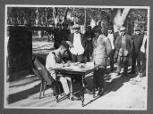Civis do sexo masculino junto a militar sentado à secretária a escrever.