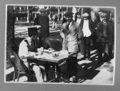 Civis do sexo masculino junto a militar sentado à secretária a escrever.